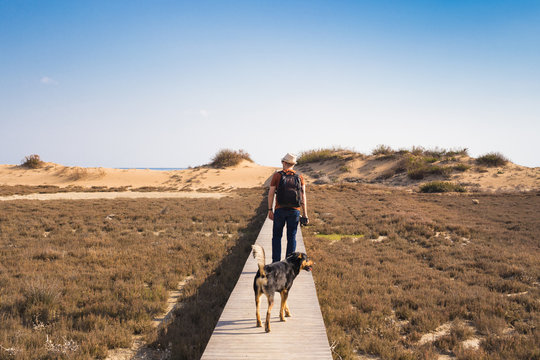 View From Behind Of A Man Walking With His Dog On A Road Leading Through Beautiful Landscape