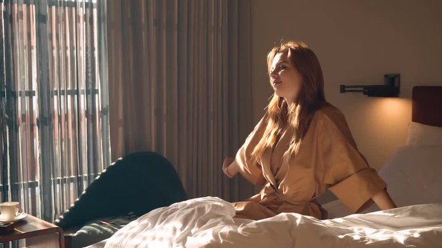 Young caucasian woman in brown beige robe streches out hands sitting on bed in the morning, looks playfully. Shot with Sony a7s and Atomos Ninja Flame.