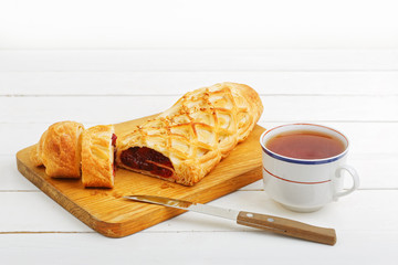 Homemade cherry strudel sprinkled with crushed nuts lying on a cutting board near a kitchen knife and cup of tea on a white wooden table.
