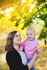 A young mother holding a small son in her arms and standing in a park near the yellow maple in the fall.