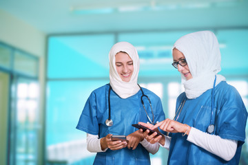 Two female middle eastern doctors in blue medical uniform standing in a hospital hallway, looking at their smart phones, smiling