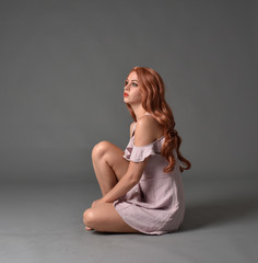 Fototapeta premium full length portrait of a pretty girl with long copper hair, wearing a simple purple dress. seated pose against a grey background.