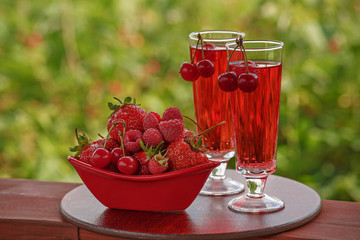 Two glasses filled with a red drink and a bowl of various berries red. Red fruit and soda water on green background with bokeh. plenty of space for text. photo of red and green colours.