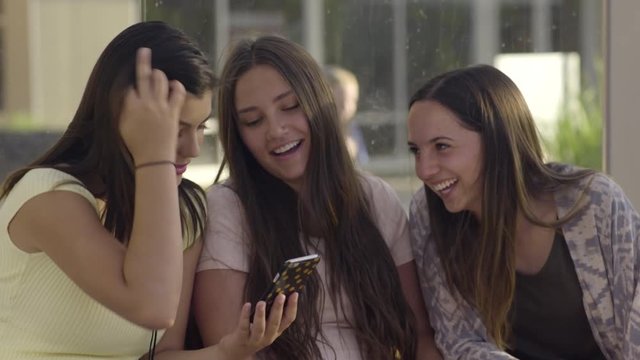 Group Of Friends Wait For Bus, Hispanic Young Woman Shows Her Friends Something Funny On Her Smartphone, They Laugh 
