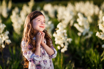 A portrait of a cute little girlwith long hair in outside at sunset in the field of white yucca flowers having fun with raised hands up
