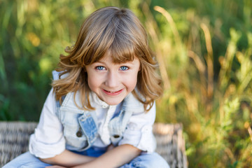 A portrait of a cute little boy with blue eyes and long blond hair in a jeans west sitting on a basket outside at sunset an having fun