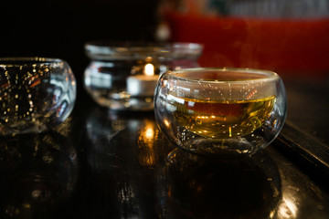 Two glass cups of tee stay on a dark black wood table. Behind of caps a beautifull candle under a glass cover. Everything is in deep dark colors under a warm light. Close up picture.