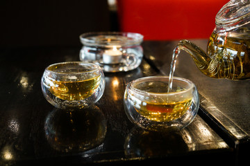 Two glass cups of herbal tee stay on a dark black wood table. Everything is in deep dark colors under a warm light. Romantic time. Water fills the cups from teapot.