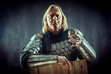 Powerful blond knight with the glass of wine on the dark background.