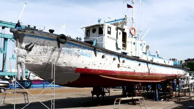 Worker paints metal of old rusty ship at shipyard in port of Moscow. Process of repair and reconstruction of sea vessel. Outdoor work. Technology of manual painting boats. Industry of water transport.