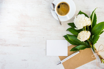 Top view of an empty greeting card, kraft envelope, peonies bouquet and a cup of coffee over white wooden rustic background.