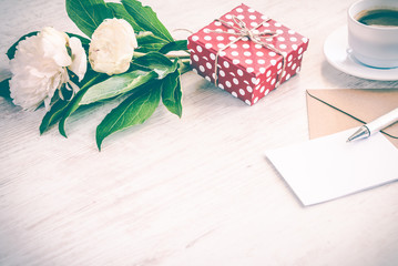 Red dotted gift box, kraft envelope and empty greeting card, coffee cup and peony flowers bouquet over white wood background. Copy space. Vintage effect.