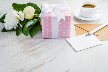 Pink dotted gift box, empty greeting card, kraft envelope, peonies bouquet and coffee cup over white wooden rustic table. Romantic holiday and event concept.
