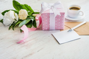 Pink dotted gift box, empty greeting card, kraft envelope, peonies bouquet and coffee cup over white wooden rustic table. Romantic holiday and event concept.