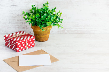 Red dotted gift box, empty card, kraft envelope and a green flower in a rustic ceramic pot. White wooden background, copy space.