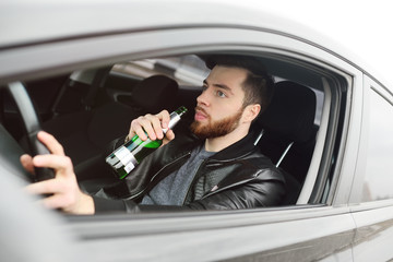 A young handsome bearded man with a bottle of beer or a low-alcohol drink at the wheel of a car. Driving in a state of intoxication