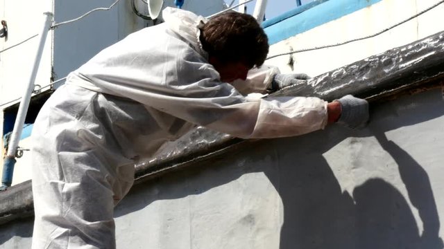 Worker paints with brush metal of rusty ship at shipyard in port of Moscow. Process of repair of sea vessel. Outdoor work. Technology of manual painting boats. Industry of water transport.