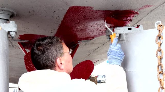 Worker paints metal of ship propeller red color at shipyard in port of Moscow. Repair and reconstruction of sea vessel. Outdoor work. Technology of manual painting boats. Industry of water transport.