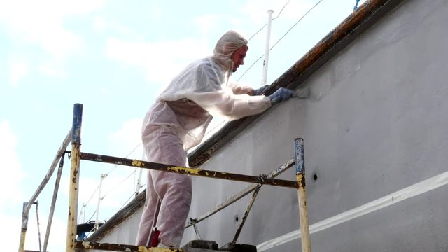 Worker paints with brush metal front of ship at shipyard in port of Moscow. Repair and reconstruction of sea vessel. Outdoor work. Technology of manual painting boats. Industry of water transport.