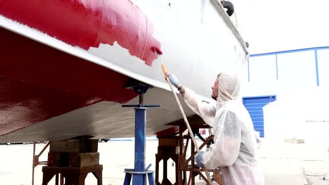 Worker paints red color metal of old rusty ship at shipyard in port of Moscow.Process of repair and reconstruction of sea vessel. Technology of manual painting boats. Industry of water transport.
