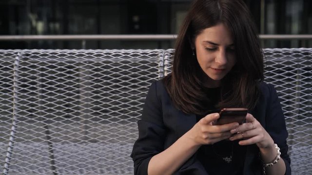 Front view of a young woman with long dark hair sitting on a rooftop in Madrid during a sunset. She is web sufring from her smartphone. Handheld real time medium shot