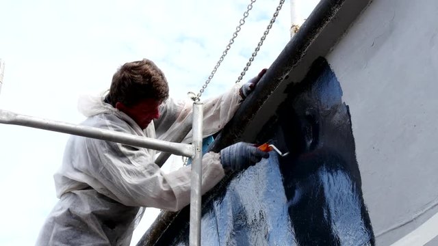 Worker paints metal of old rusty ship at shipyard in port of Moscow. Process of repair and reconstruction of sea vessel. Outdoor work. Technology of manual painting boats. Industry of water transport.