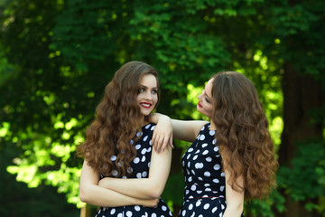 Fototapeta premium Two beautiful smiling girl sisters twins for a walk in summer park on a background of greenery with makeup and hairstyle