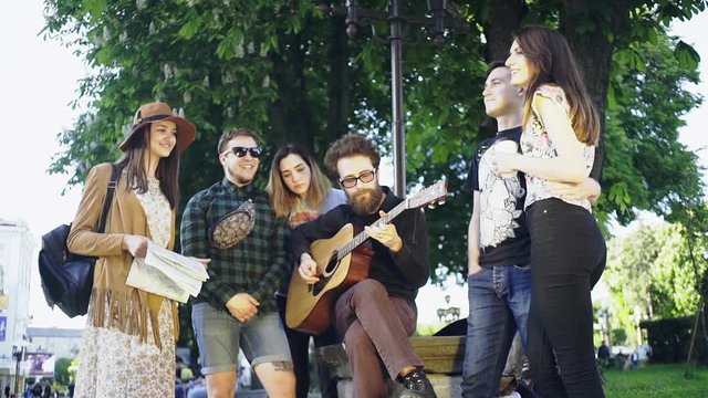 Group of young people hangout at the city street. They sitting on bench, singing and playing guitar.