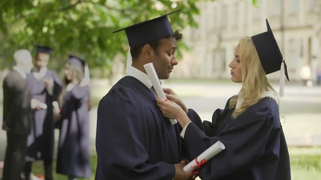 Graduates in academic regalia talking, girl fixing tie, guy kissing her on cheek