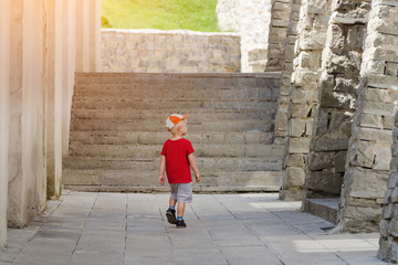 Fototapeta na wymiar Little boy walking alone among the ancient walls, sunlight. Back view