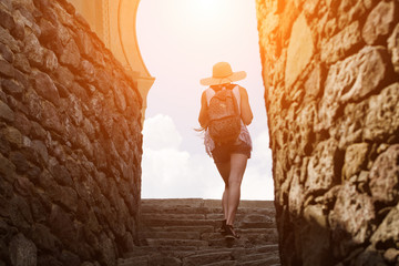 Girl in a hat with a backpack climbs the stairs of the fortress, sunlight. Back view