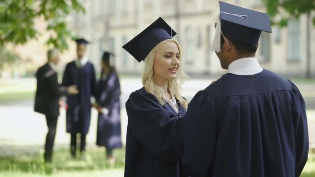Graduate female student talking to male graduate, touching hat, happy friends