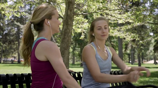 Happy Young Women Listen To Music And Stretch On A Bench, Then They Get Up And Jog (Steadicam Follows Along) 