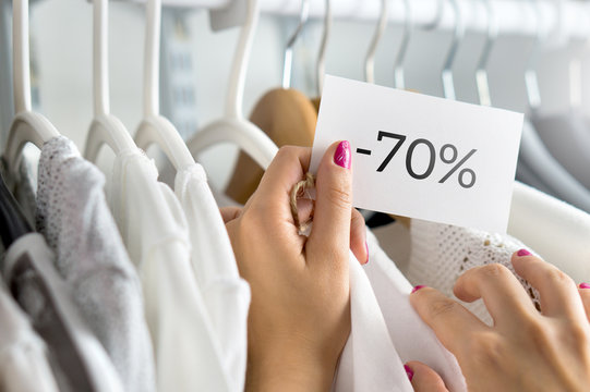 Female customer browsing clothes in a shop. Woman shopping for fashion offer and deal. Holding price tag with minus seventy percent or 70% sale, bargain and reduced cheap prices in clothing store.