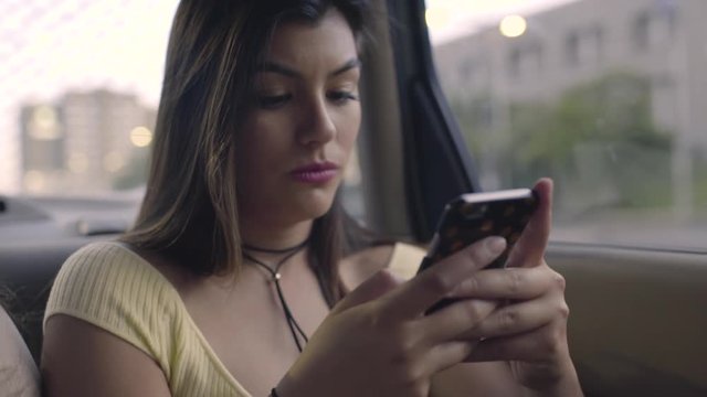 Friends Use A Rideshare, Closeup Of Hispanic Young Woman Using Her Smartphone In Back Seat Of Moving Car