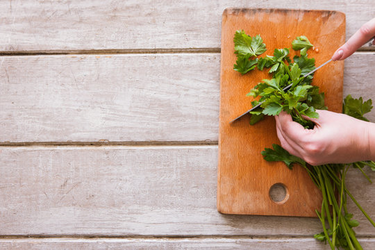 Woman cuts parsley on wooden desk with steel knife. Herbs as healthy and organic food, top view picture, free space nearby