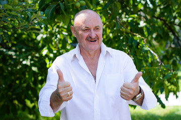 Successful excited elderly man in white shirt showing ok sign while standing in a garden background. Happy, successful, positive middle aged man showing thumb up