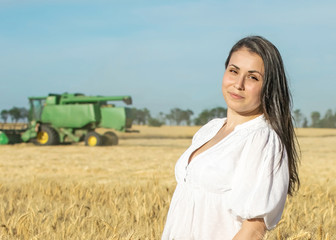 Pretty female standing in wheat field during harvesting. Harvester in the back blurry background