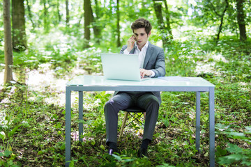Fototapeta premium Young handsome business man sitting at office desk in the green park or forest and work at laptop while talk at phone with costumer. Business concept.