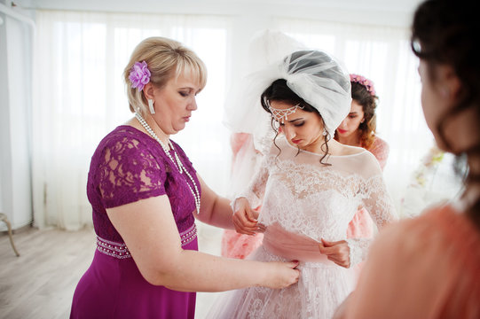 Helpful bridesmaids and mother helping bride to tie the dress up in big light room.
