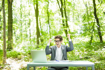 Fototapeta premium Portrait of young handsome business man in suit working at laptop at office table with raised hands achived goals in green forest park. Business concept.