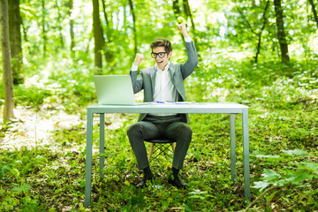 Fototapeta premium Portrait of young handsome succesful manager in suit working at laptop at office table with raised hands in green forest park. Business concept.