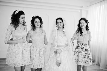 Fantastic bride posing in wedding dress with helpful pretty bridesmaids in big white room. Black and white photo.