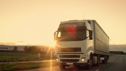 Front-View of Semi-Truck with Cargo Trailer Driving on a Highway. He's Speeding Through Industrial Warehouse Area with Sunset in the Background.