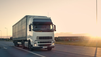 Plakat Front-View of Semi-Truck with Cargo Trailer Driving on a Highway. He's Speeding Through Industrial Warehouse Area with Sunset in the Background.