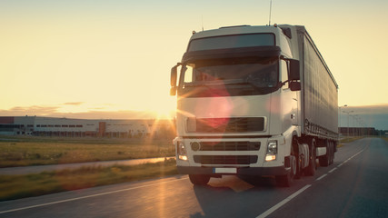 Naklejka premium Front-View of Semi-Truck with Cargo Trailer Driving on a Highway. He's Speeding Through Industrial Warehouse Area with Sunset in the Background.