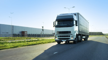 Front-View of Semi Truck with Cargo Trailer Driving on a Highway. He's Speeding Through Industrial Warehouse Area while Sun Shines and no other Vehicles are on the Road.
