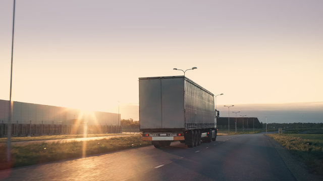 Shot of a Semi-Truck with Cargo Trailer Moving on a Highway. White Truck Drives Through Industrial Warehouse Area on an Empty Road with Sun Shining in the Background.