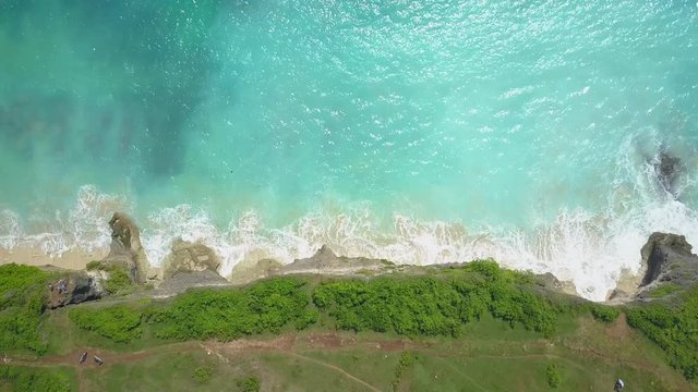 AERIAL TOP DOWN: Gentle foamy whitewater ocean waves splashing against cliffy shore washing white sandy beach in sunny Bali island. Lush overgrown rocky wall rising above the crystal clear emerald sea