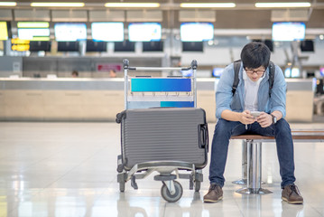 young asian guy using smartphone while waiting for check in and drop his suitcase luggage at airline check-in counter of international airport terminal